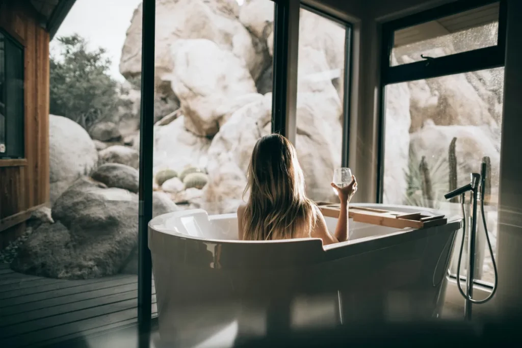 A girl is taking a bath in the bathtub holding a wine glass in her hand and there is a bathtub tray table in front of her