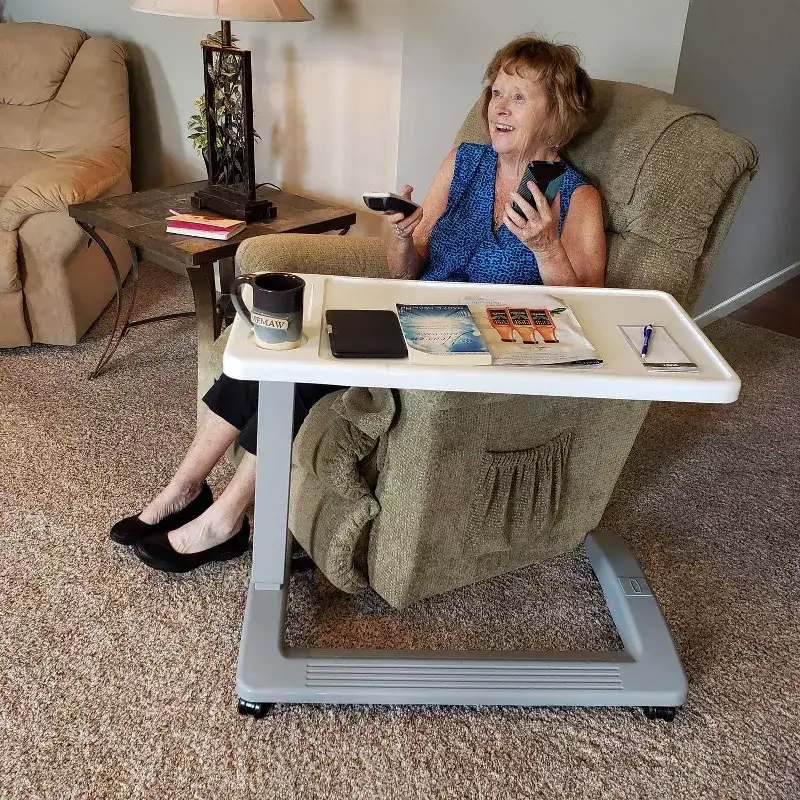 An old woman is sitting on the sofa watching TV There are water glasses books and pens on the TV tray table next to her